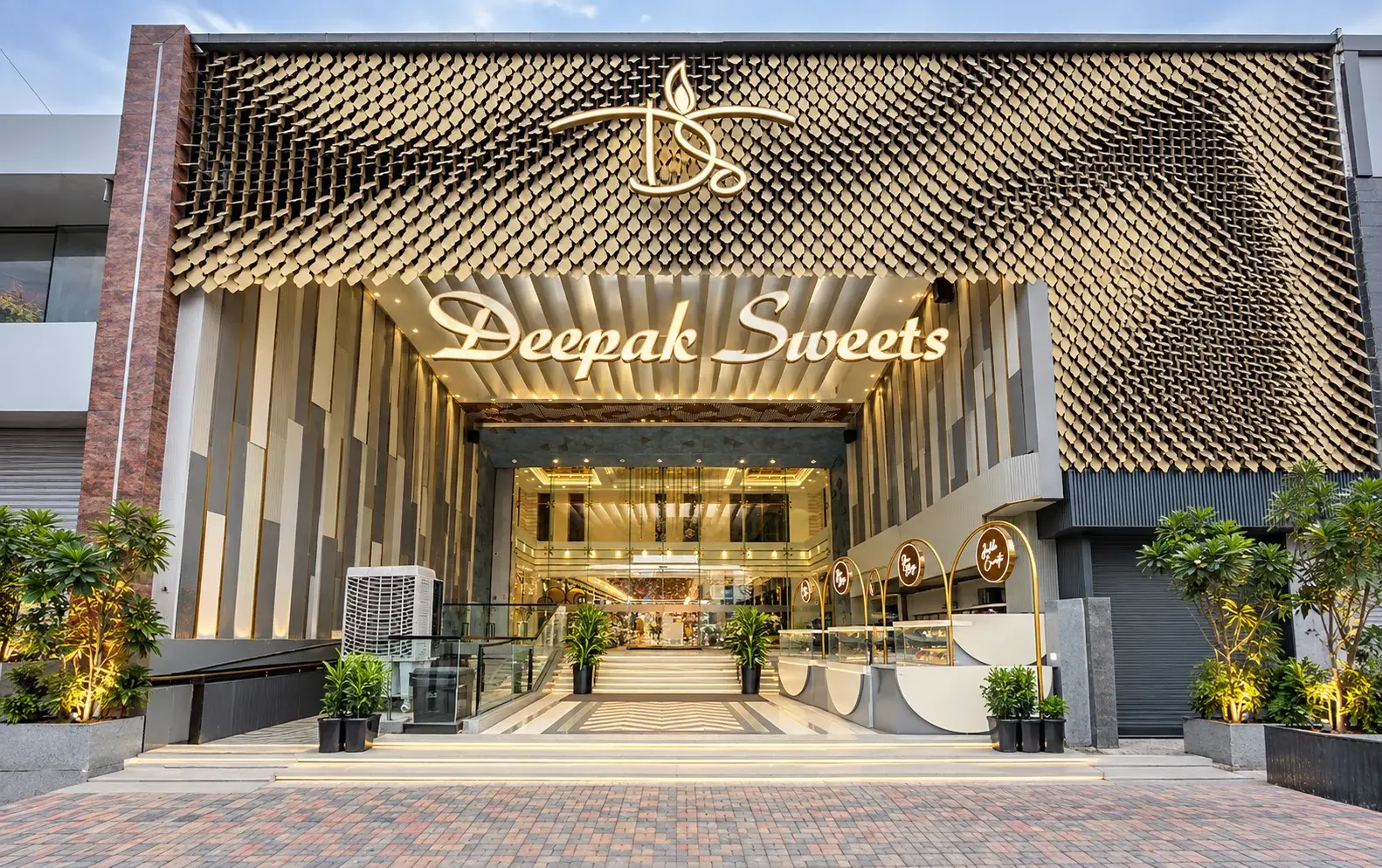 The Deepak Sweets storefront: an illuminated Deepak Sweets sign beneath a gold scaled facade, with steps leading to a glass entrance